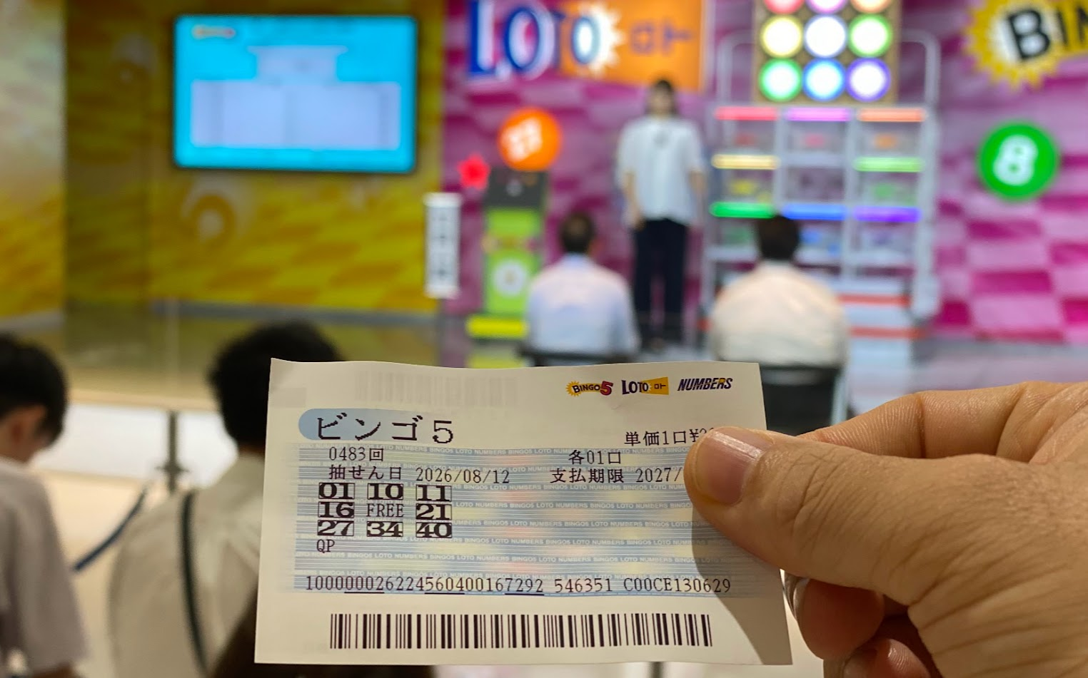 購入したビンゴ5の宝くじを手に東京宝くじドリーム館の抽せんライブを見る様子